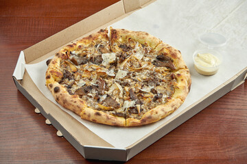 Photo for pizza delivery. Pizza with different mushrooms, parmesan and white sauce in a cardboard box with sauce for the sides on a wooden table