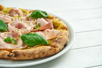 Appetizing homemade pizza with mortadella sausage, pistachios, basil and melted cheese on a white plate on a white wooden background
