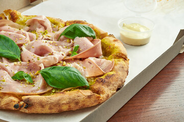 Photo for pizza delivery. Pizza with mortadella sausage, pistachios, basil and melted cheese in a cardboard box with side sauce on a wooden table