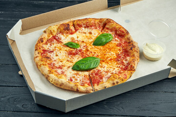 Photo for pizza delivery. Pizza margherita with tomatoes, basil and mozzarella in a cardboard box with sauce for sides on a wooden table