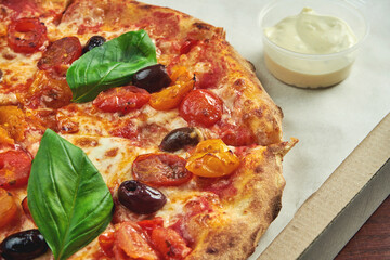 Photo for pizza delivery. Margarita pizza with cherry tomatoes, basil and mozzarella in a cardboard box with side sauce on a wooden table
