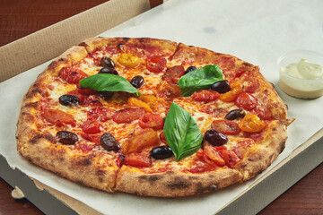 Photo for pizza delivery. Margarita pizza with cherry tomatoes, basil and mozzarella in a cardboard box with side sauce on a wooden table