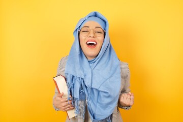 Close up portrait of stylish muslim woman wearing hijab raising fists up isolated on shine background big eyes and wide open mouth.