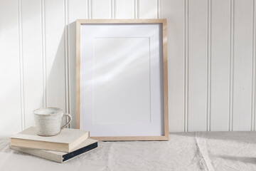 Empty wooden frame mockup on beige linen tablecloth background. White wooden wall paneling background. Artistic still life with cup of coffee and books in sunlight. Scandinavian interior. Art concept.