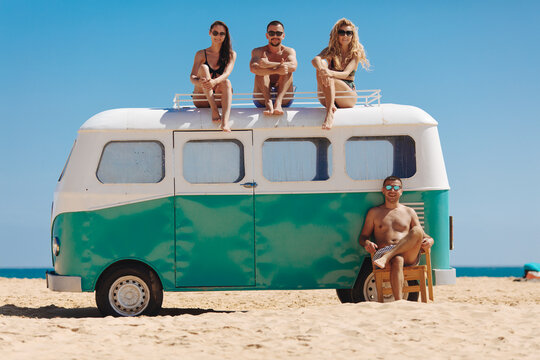 Group of four smiling travelers in sunglasses and swimsuits taking fun near retro hippie car on beach. Happy friends spending time together on Hainan island. Beautiful place to visit.