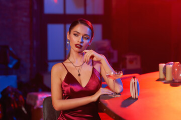 Elegant brunette girl with pony tail hairstyle and perfect makeup with glass of sparkling wine relaxing near bar counter at party. Colorful red and blue light