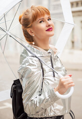 Outdoors lifestyle beauty close up portrait of ginger hair girl under transparent umbrella. Smiling and walking at the city. Wine lips. Perfect eyebrows. Blue eyes.  Rainy weather season concept.