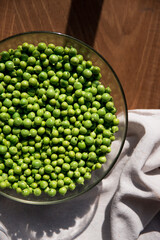 Crystal bowl full of green peas on a wooden table in a sunny day. In the table there is a linen cloth. Rustic style. Top view.