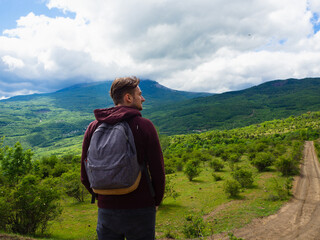 A young guy in a red hoodie and gray backpack travels in the mountains among green trees and clouds.