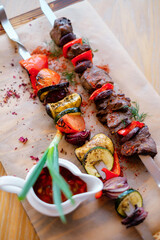 Fried meat on a skewer with potatoes and vegetables. Grilled BBQ decorated with vegetables and sauce on wooden table and parchment paper.