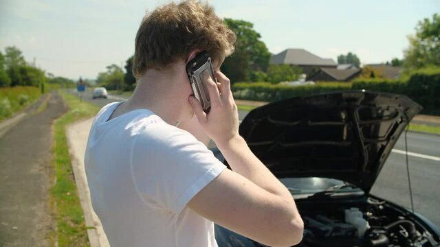 Young man on his mobile phone contacting help for broken-down vehicle, car break down on main road in background cars drive past whilst gentlemen has his bonnet open showing his broken engine