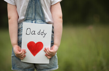 Father's Day. Close-up  of a child girl hands holding a card with congratulation for dad outdoor. Back view. Copyspace