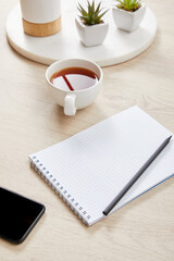 green plants, cup of tea and blank notebook with pencil near smartphone on wooden surface