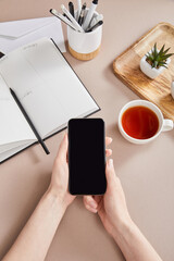 female hands with smartphone near green plants on wooden board, cup of tea, planner with pencils and envelope on beige surface
