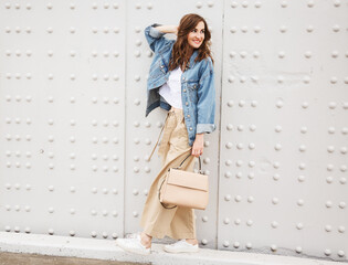 Outdoors fashion portrait of trendy pretty girl posing on the white wall background. Smiling and walking on the city. Going shopping. Wearing stylish wide jean jacket, beige slacks. Positive emotiones