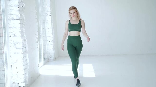 young sexy fitness girl in leggings and a T-shirt with a beautiful athletic body goes to the camera with her hands up. Blonde woman posing at camera. Slow motion. White studio background