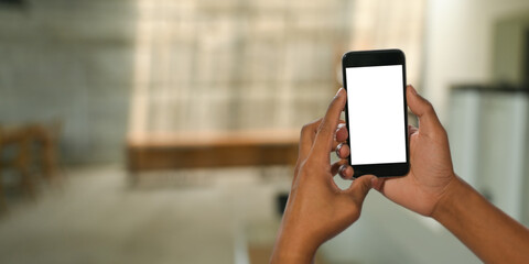 Cropped image hands holding cropped black smartphone with white blank screen over blurred comfortable sitting room as background.