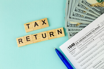 Tax return concept. Still life of US individual Income Tax Form 1040 pen and inscription text on wooden blocks with several 100 dollar banknotes on a blue pastel background.