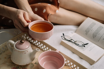 A woman holds a cup of hot tea on the bed at home and reads a book, home leisure in coziness and comfort.