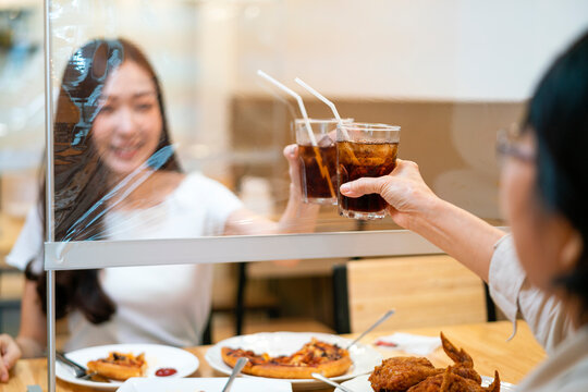 Asian Beautiful Female Clinking A Glass Of Water With Her Mother Who Siting Separate And Keep Distance With Table Plastic Shield Partition In Restaurant, New Normal And Social Distancing Concept