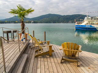 Umbrella, table and chairs placed on deck that borders calm Wörthersee in Austria with bow of ferry shown on water in the background. 