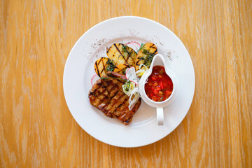 Fried  barbeque meat on a white plate with potatoes and vegetables. Grilled BBQ decorated with vegetables and sauce on wooden table and parchment paper.