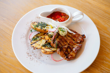 Grilled  barbeque meat on a white plate with potatoes and vegetables. Grilled BBQ decorated with vegetables and sauce on wooden table and parchment paper.