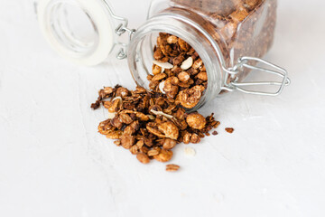 A jar of home made granola spilling on to a white table top. This granola is made from roasted oats with chocolate,cinnamon and almonds for a delicious breakfast. Copy space.