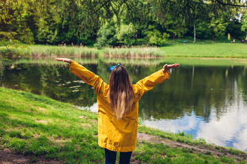 Fototapeta na wymiar Happy woman in yellow raincoat raising arms feeling free walking in summer park. Girl admires lake landscape view