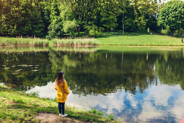 Fototapeta na wymiar Lonely woman in yellow raincoat walking in summer park. Girl admires lake landscape view after rain