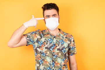 Unhappy man wearing hawaiian shirt and medical mask curves lips and makes suicide gesture, tired of everything, shoots in temple with hand, isolated against studio background.