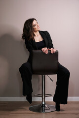 Full length studio portrait of young business woman in black suit sitting in chair on grey background