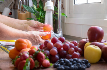 Woman's hands washing two apricots under the water flush, assortment of fresh fruit in kitchen, bright light from window