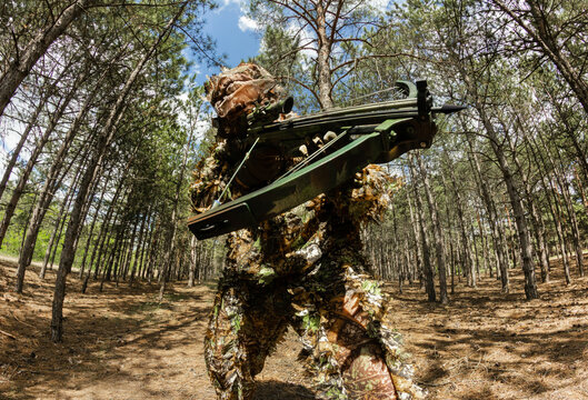 Forest sniper hunter in camouflage leaf suit posing and aiming with crossbow.