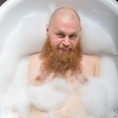 Fototapeta premium Portrait of a bald man fooling around in the foam bath. Top view on guy having fun in the bathroom.