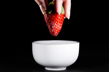 Strawberry with sugar held by woman's fingers on black background and white ceramic bowl on black background