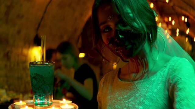 Young woman with scary make up holding a drink and candles. Stock footage. A girl waitress with a creepy face of a zombie during Halloween celebration at the night club.