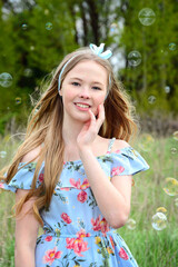 A smiling blonde girl in a blue dress is standing against a background of soap bubbles with hair...