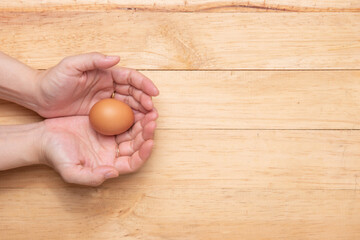 Top view of two hands holding a raw chicken egg on a light brown wooden background, copy space