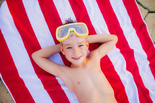 Little Boy In Diving Goggles Lying On Red And White Beach Towel And Smiling