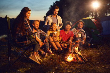 A group of people sitting by the bonfire next to the tent at night in the summer in autumn.