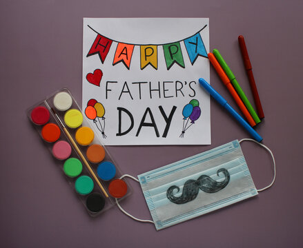 Greeting card Happy Father’s Day, color paints and markers and protective mask with mustache drawn by a child as a gift for dad. Violet background. Father’s day in the context of coronavirus pandemic