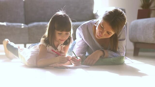 Mother and small daughter lying on warm floor in living room using colored pencils drawing on album paper, elder sister or nanny helps teaches kid girl, leisure activities, talent development concept