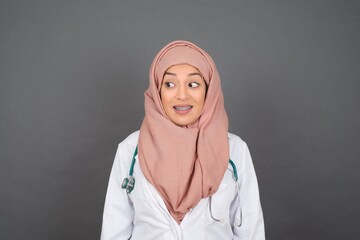 Oops! Portrait of muslim doctor female clenches teeth and looks confusedly aside, realizes her bad mistake, poses against copy area for your advertising or promotional background.