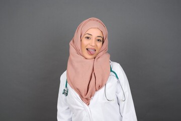 Beautiful muslim doctor woman with happy and funny face smiling and showing tongue. Wearing medical uniform and standing against gray studio background.