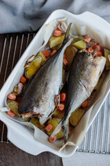 Two delicious and appetizing sea fish baked with potatoes, carrots, onions and garlic in a baking sheet on a wooden brown table. Homemade tasty and simple food. Fish cooked in the oven. 
Top view 