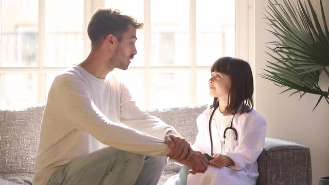 Daughter and father sitting on couch playing having fun together girl wearing white coat imagines herself to be doctor hold pen listens daddy makes notes, leisure activities, future profession concept