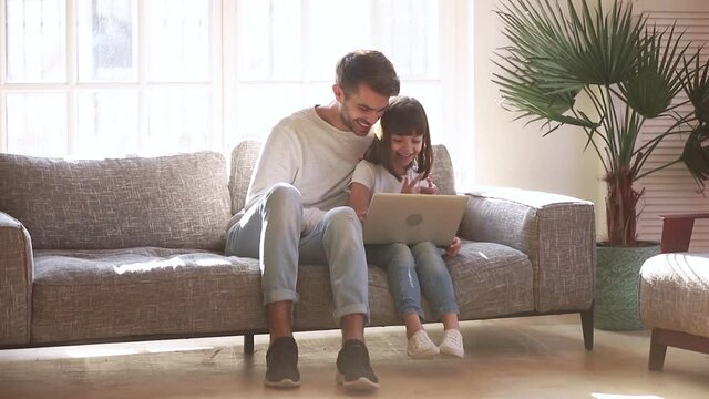 Father and daughter sit on sofa spend time having fun together fool around using laptop apps, kid make funny faces laughing, take photos on web camera, modern wireless tech leisure activities concept
