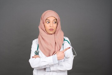 Excited surprised muslim girl wearing hijab and medical uniform points happily aside on copy space of gray background for your advertising information or promotional text.