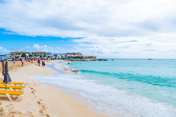 Philipsburg, Sint Maarten. Maho and Sunset Beach Bar with pristine white sand and water. Tourists swim, stand on shore ans await overhead aircraft landing at SXM Airport, next door.